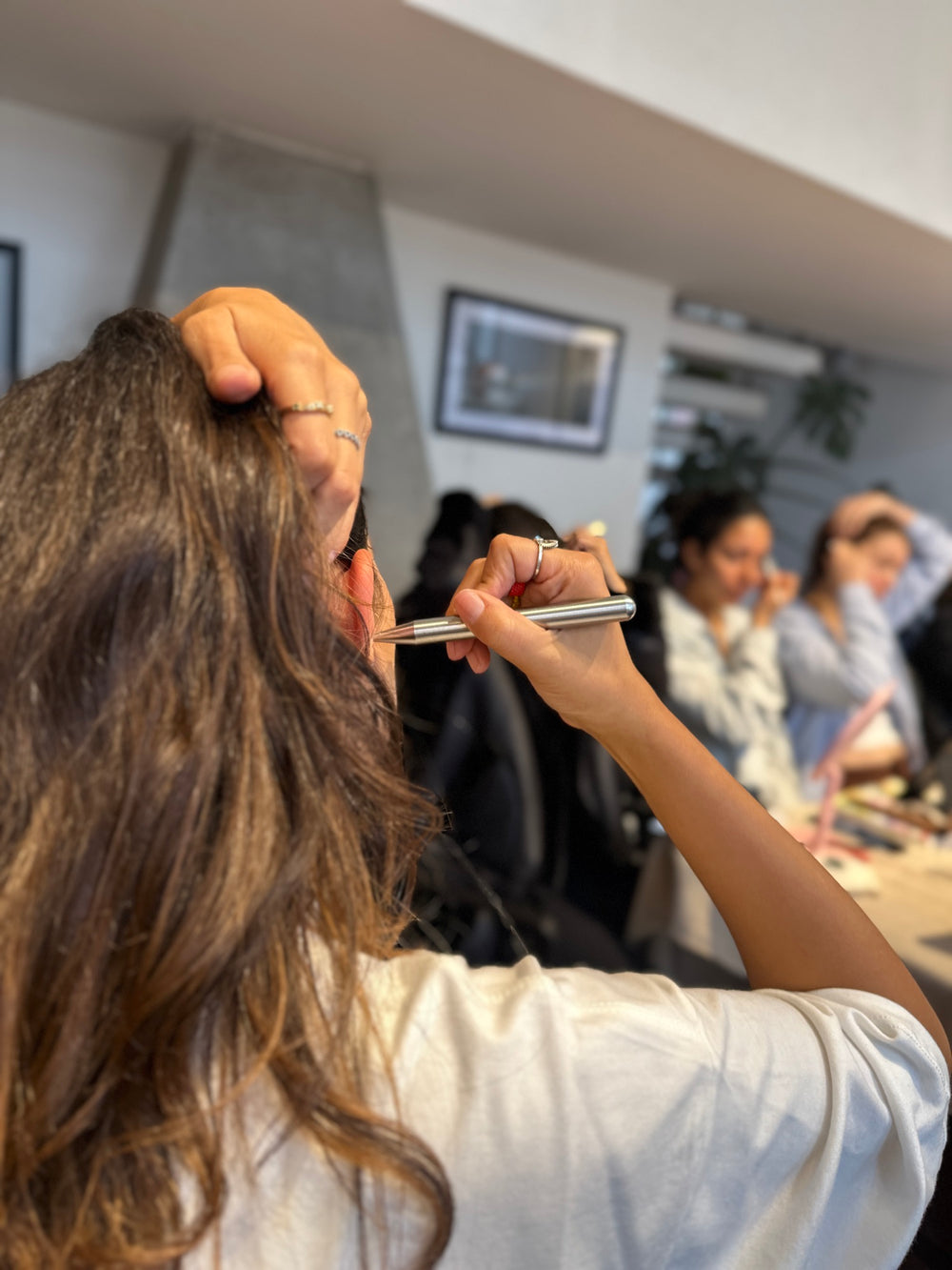De espaldas, una mujer se pasa la varita de reflexología facial por el rostro con la mano en la cabeza; al fondo, el grupo practica