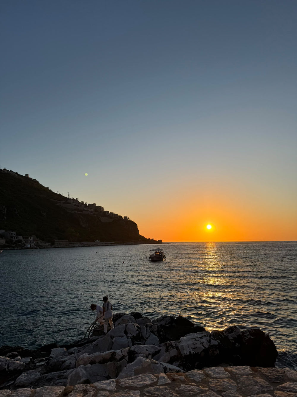 Atardecer naranja sobre el mar, con una barca y dos personas en las rocas